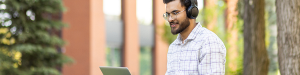 A young man with glasses and headphones uses a laptop outdoors, smiling as he works.